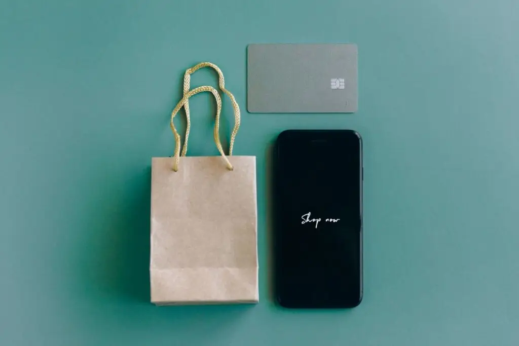 Smartphone com a mensagem compre agora ao lado de cartão de crédito e sacola de compras